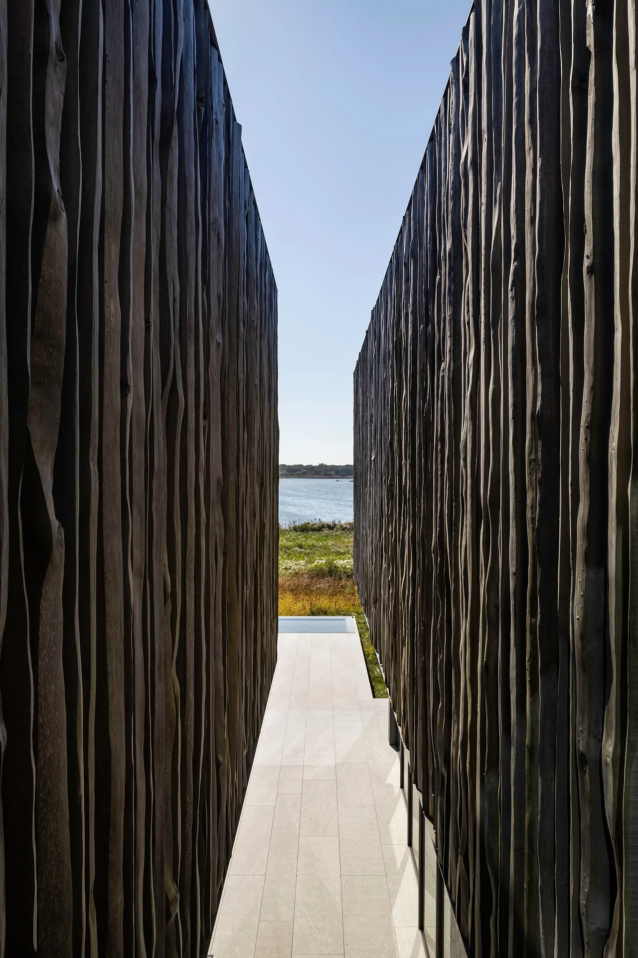 Narrow passage formed by two tall walls clad in irregular dark wood planks leading to a waterside view.