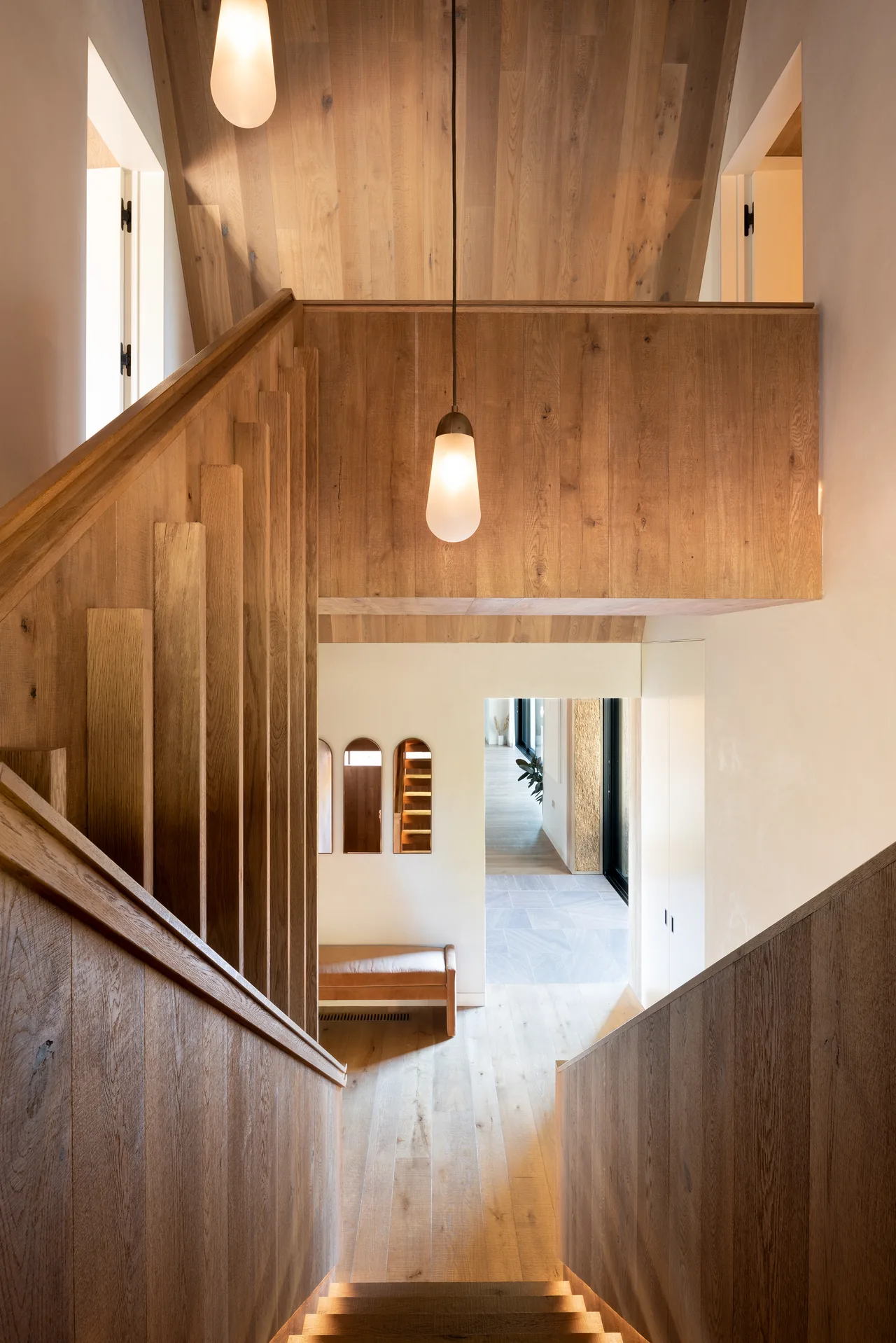 Wood-paneled staircase with a pendant light and light wood flooring leading to a hallway with mirrors and bench seating.