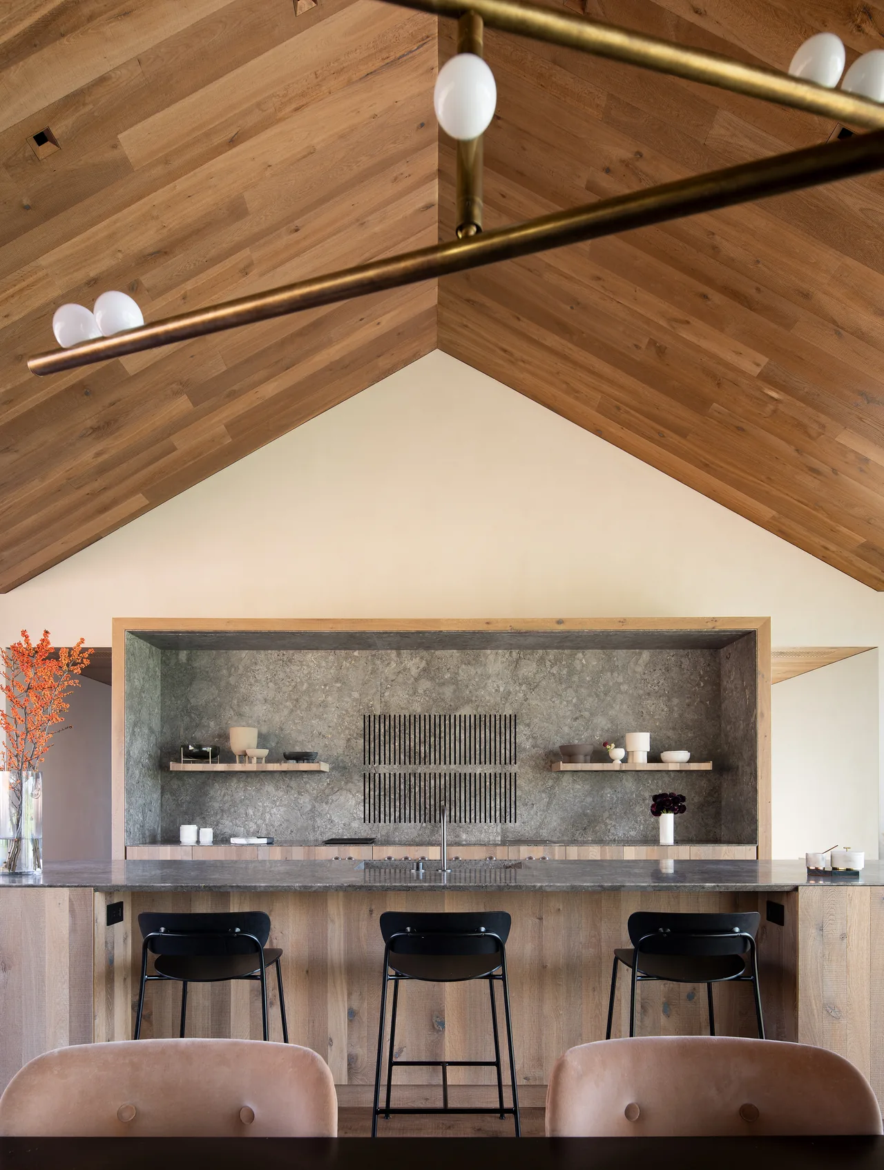Kitchen features a vaulted wood-planked ceiling, gray stone backsplash, wood cabinetry, and marble island with black stools.