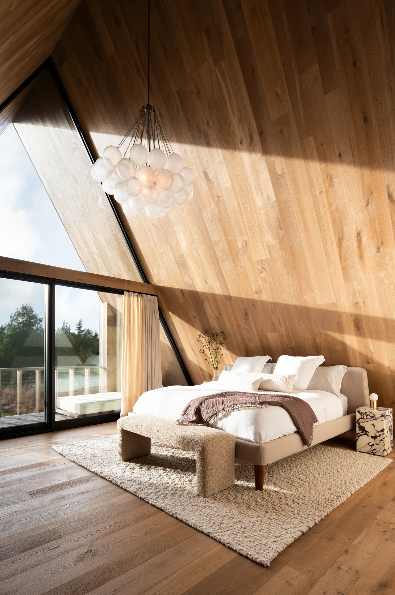 Bedroom interior with wood-paneled sloped walls and ceiling, a modern bubble chandelier, and floor-to-ceiling glazing.