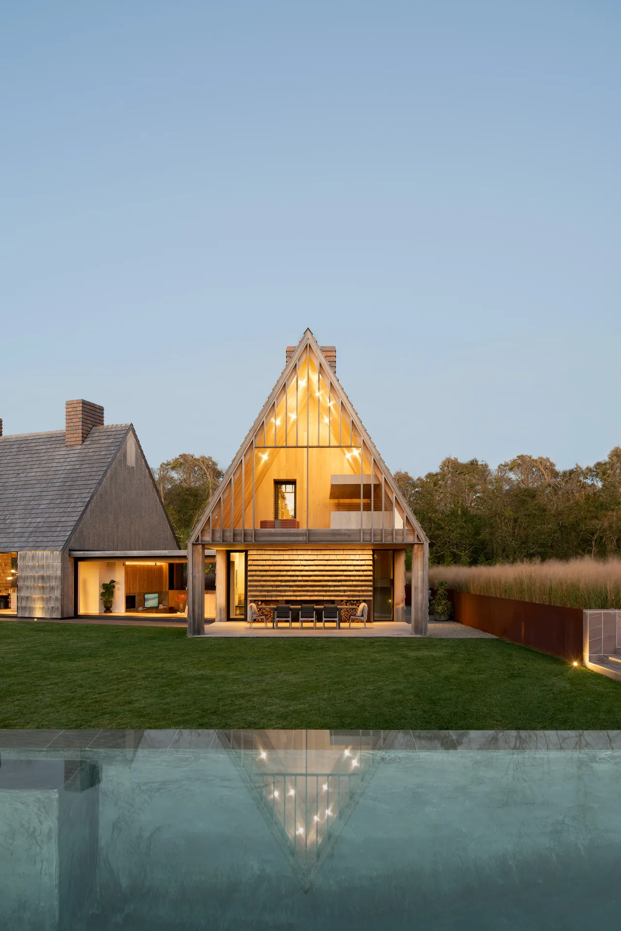 Modern steep-pitched house with extensive vertical glazing and wood paneling, viewed from the yard with pool reflection.