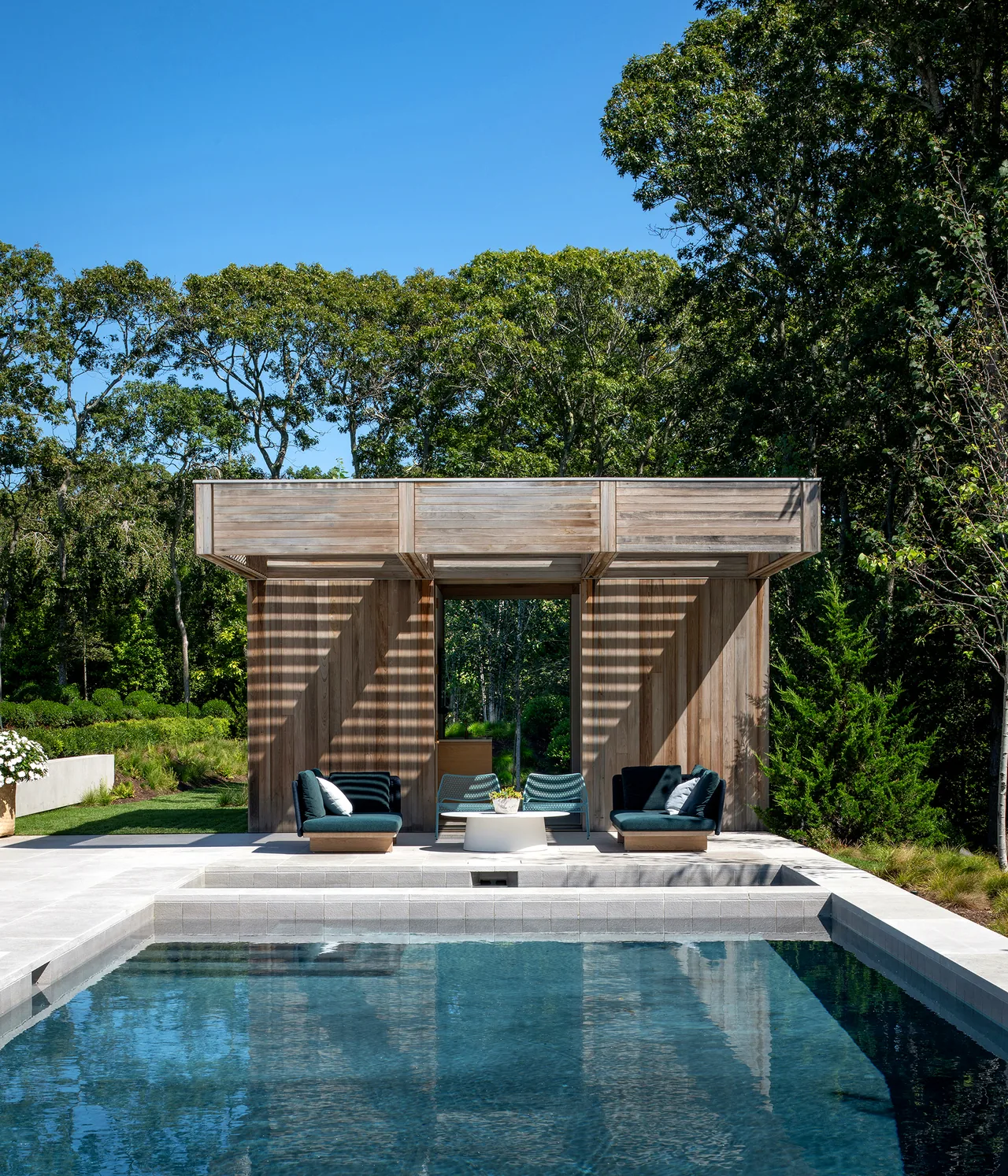 Swimming pool with wooden pergola seating area in a natural wooded setting.