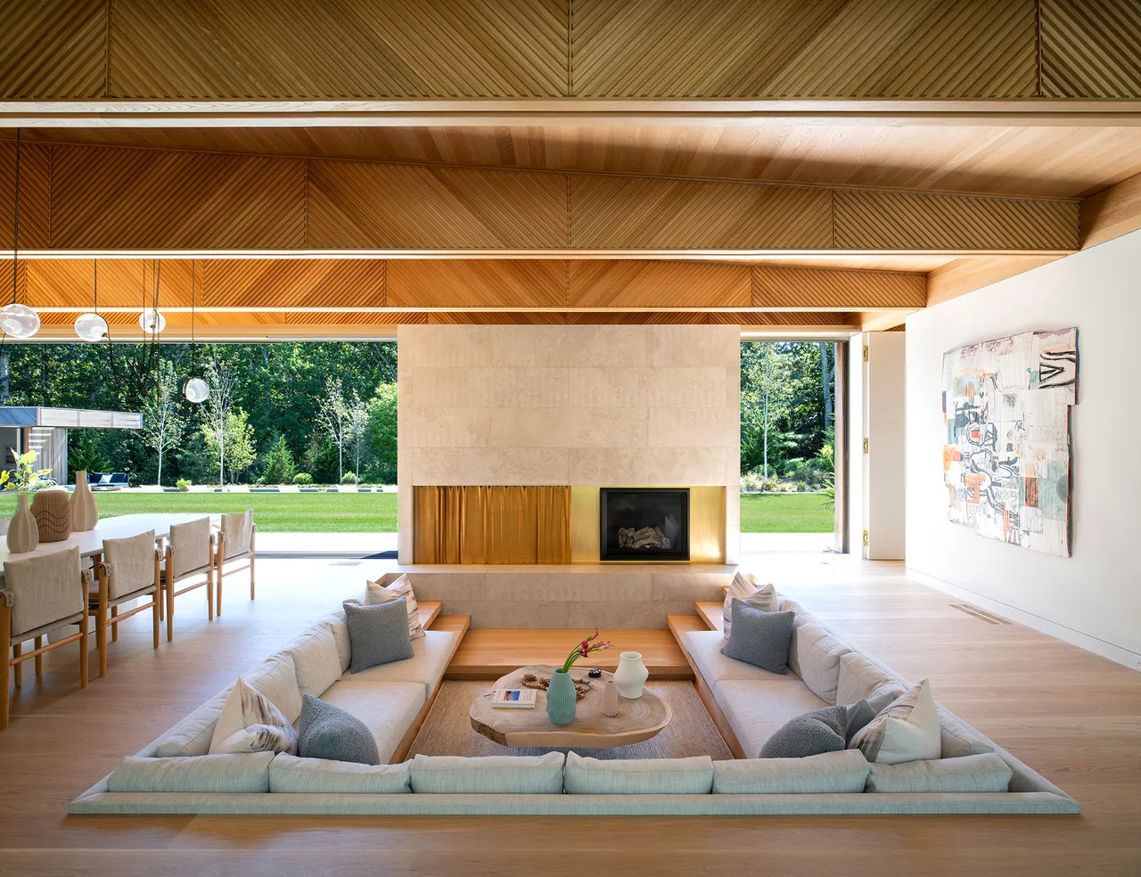 Sunken living room with a stone fireplace, wooden ceiling beams, and adjacent dining area overlooking a grassy outdoor space.
