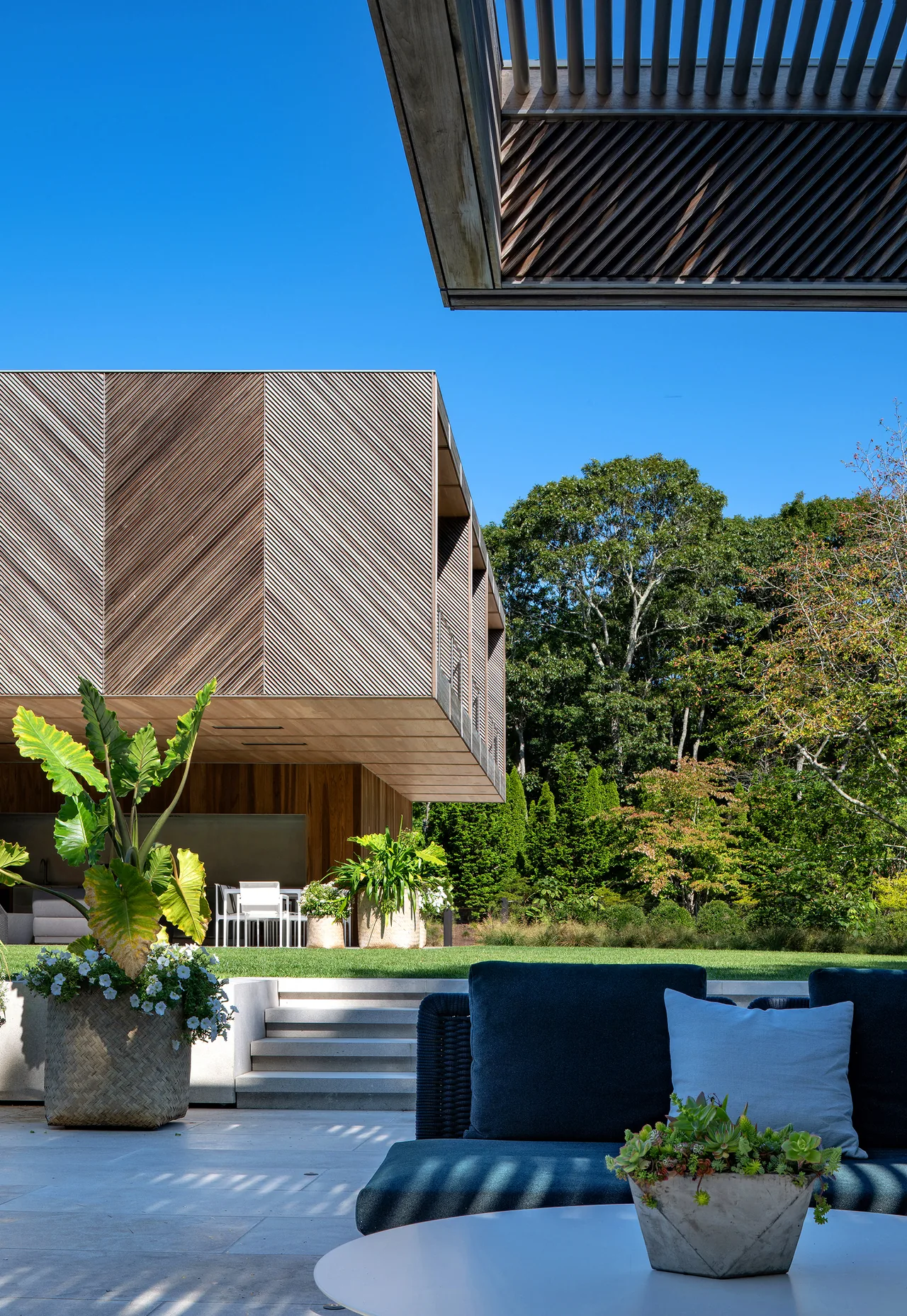 Outdoor patio area with wood-clad upper structure and woven wood detailing, surrounded by greenery under a clear blue sky.