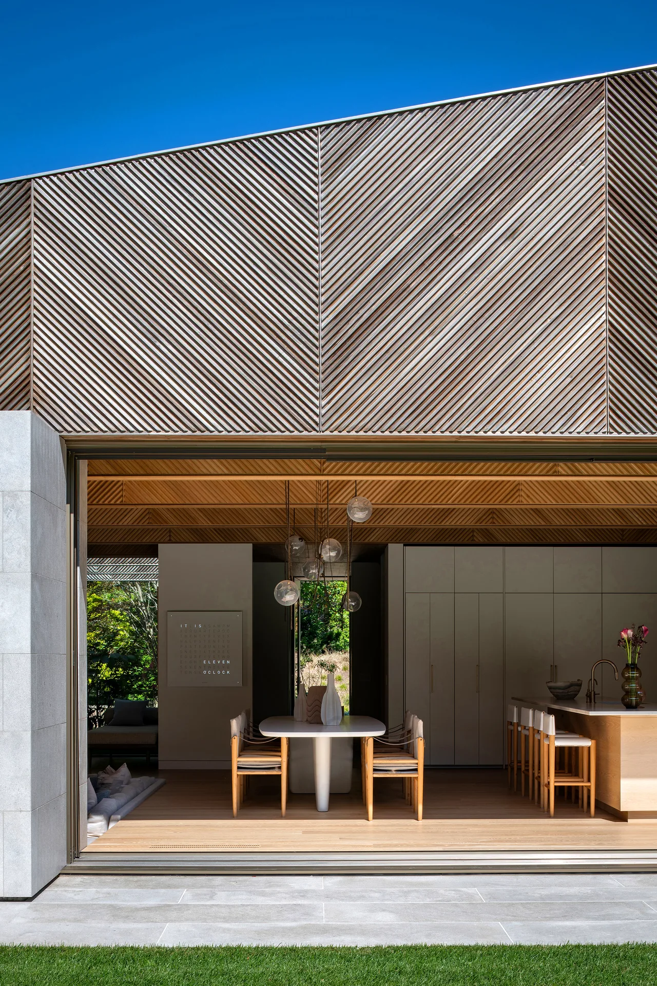 Open sliding glass doors connect the dining room and kitchen interior to the outdoor area with a chevron-patterned wooden ceiling feature above.