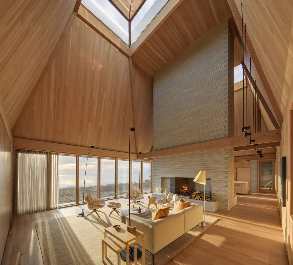 Spacious living room with vaulted wood-paneled ceiling, brick fireplace, and floor-to-ceiling glazing overlooking outdoor scenery.