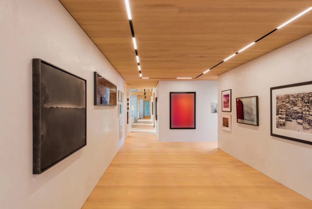 Hallway with artwork displayed on white walls, wood flooring and ceiling, and integrated linear recessed lighting.