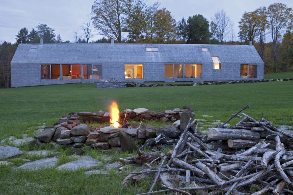 Farmhouse with wood shingle exterior and skylights, seen from the lawn with a stone fire pit in front at dusk.