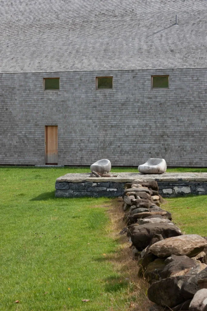 A side exterior wall of a shingled building with three small windows and a wooden door alongside a stone wall and two stone chairs.