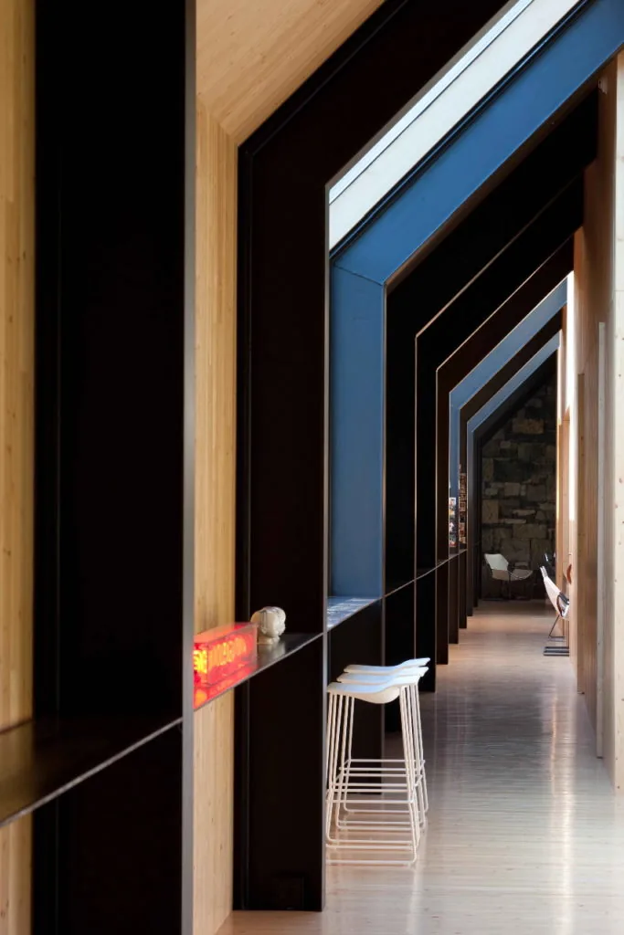 Long interior corridor with angled black structural frames and wood paneling, featuring stools and minimalistic decor.