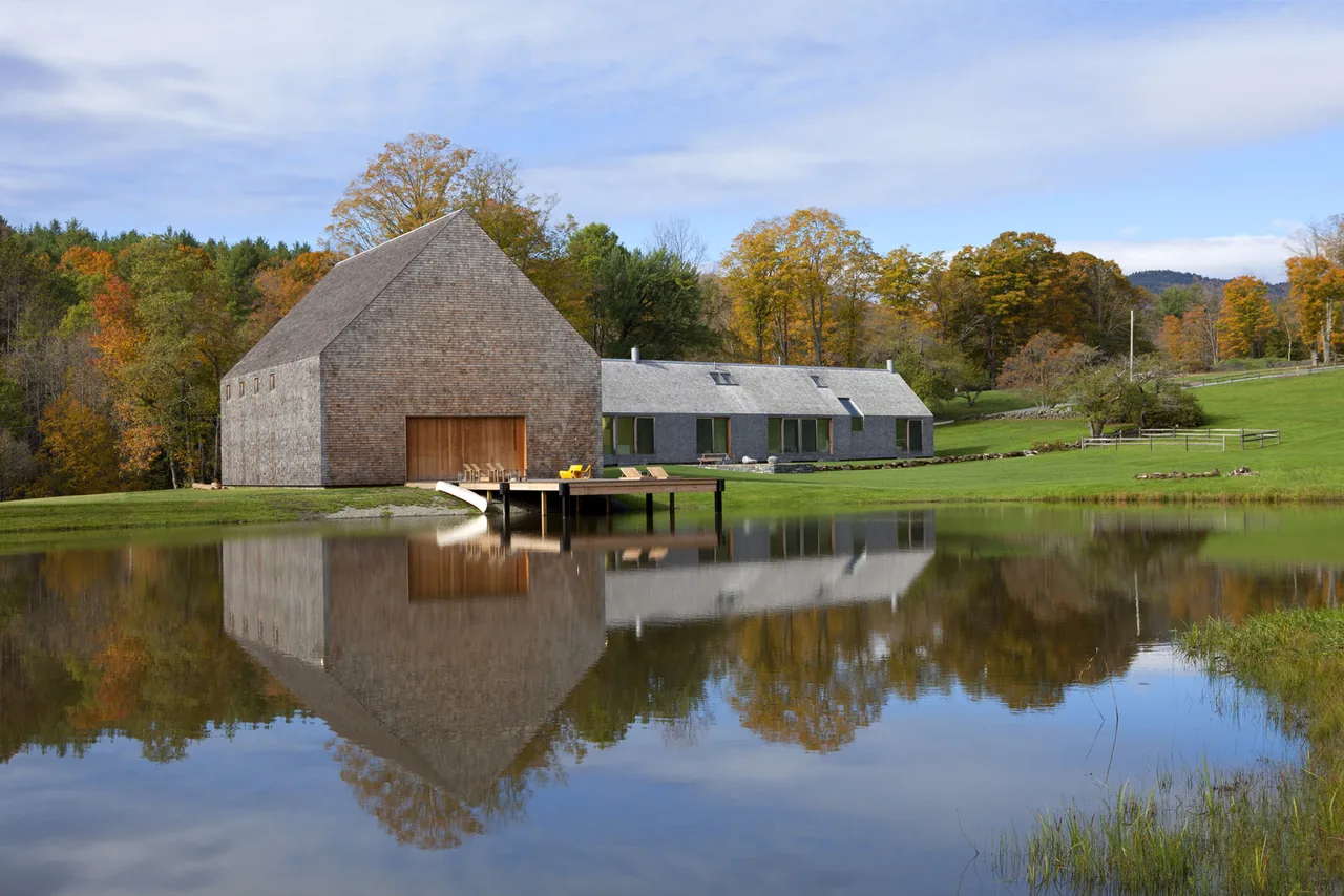 Barn-style gable-roofed building and adjacent long house beside a pond in a rural autumn landscape.