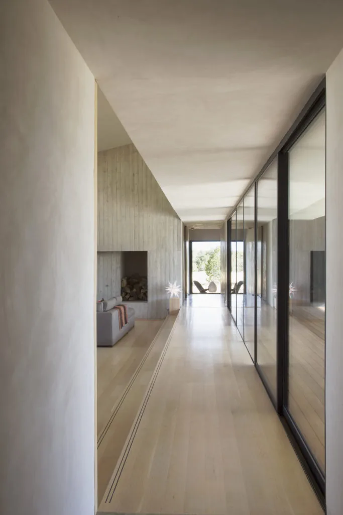 Interior hallway featuring light wood flooring and floor-to-ceiling glass doors next to a sitting area with a recessed concrete fireplace.