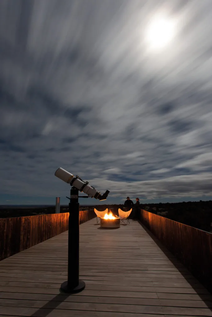 Nighttime view of the rooftop deck featuring a telescope, fire pit with chairs, and surrounding wooden railing.