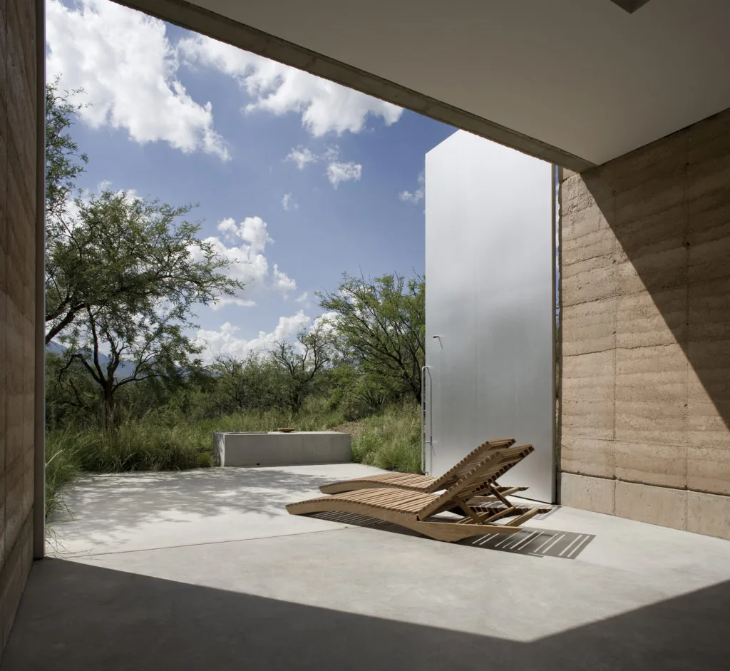 Covered patio with rammed earth walls and wooden lounge chairs opens to natural landscape under blue sky.