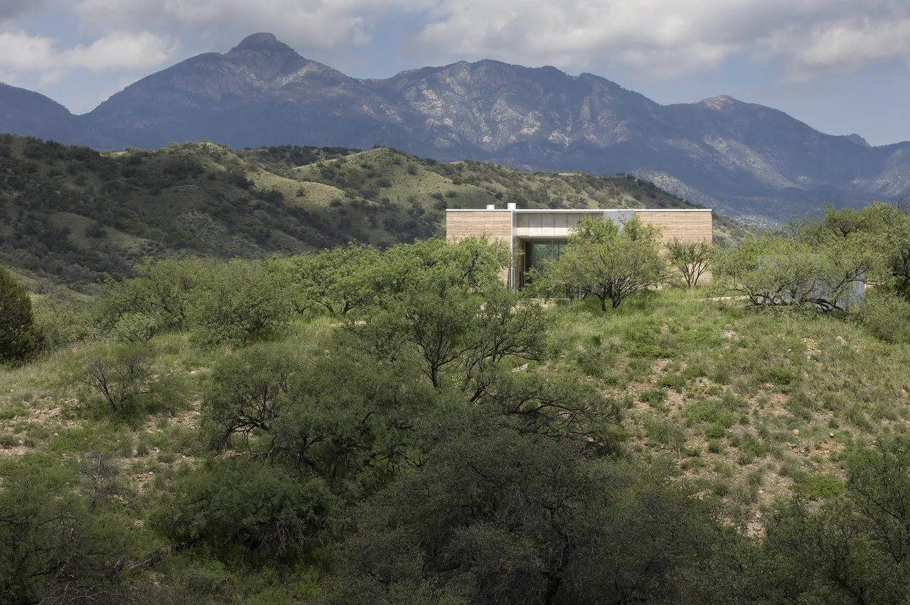 Modern single-story house situated on a hillside with natural vegetation and mountains in the background.
