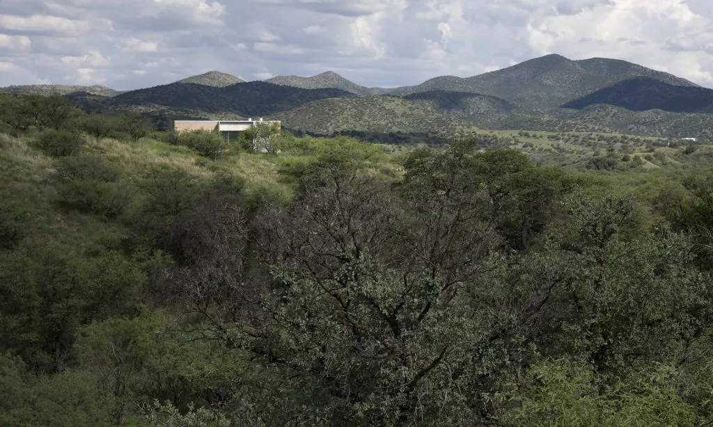 Adobe Canyon House sits amid dense greenery with mountainous terrain in the distance under a partly cloudy sky.