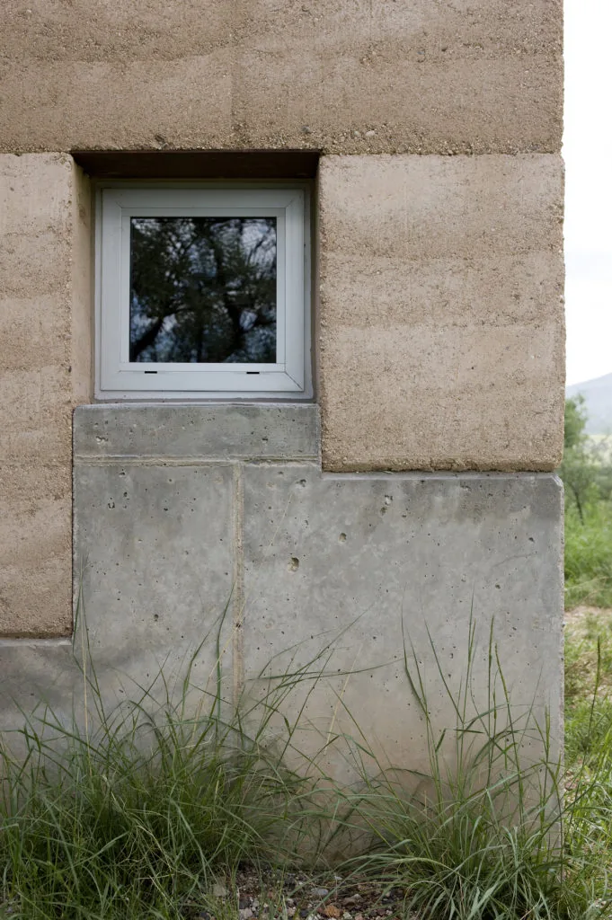 Window detail showing rammed earth walls above and concrete base below with vegetation at ground level.