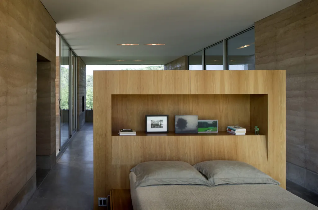The bedroom in Adobe Canyon House features a wood headboard with shelving, rammed earth walls, and views through floor-to-ceiling glass.