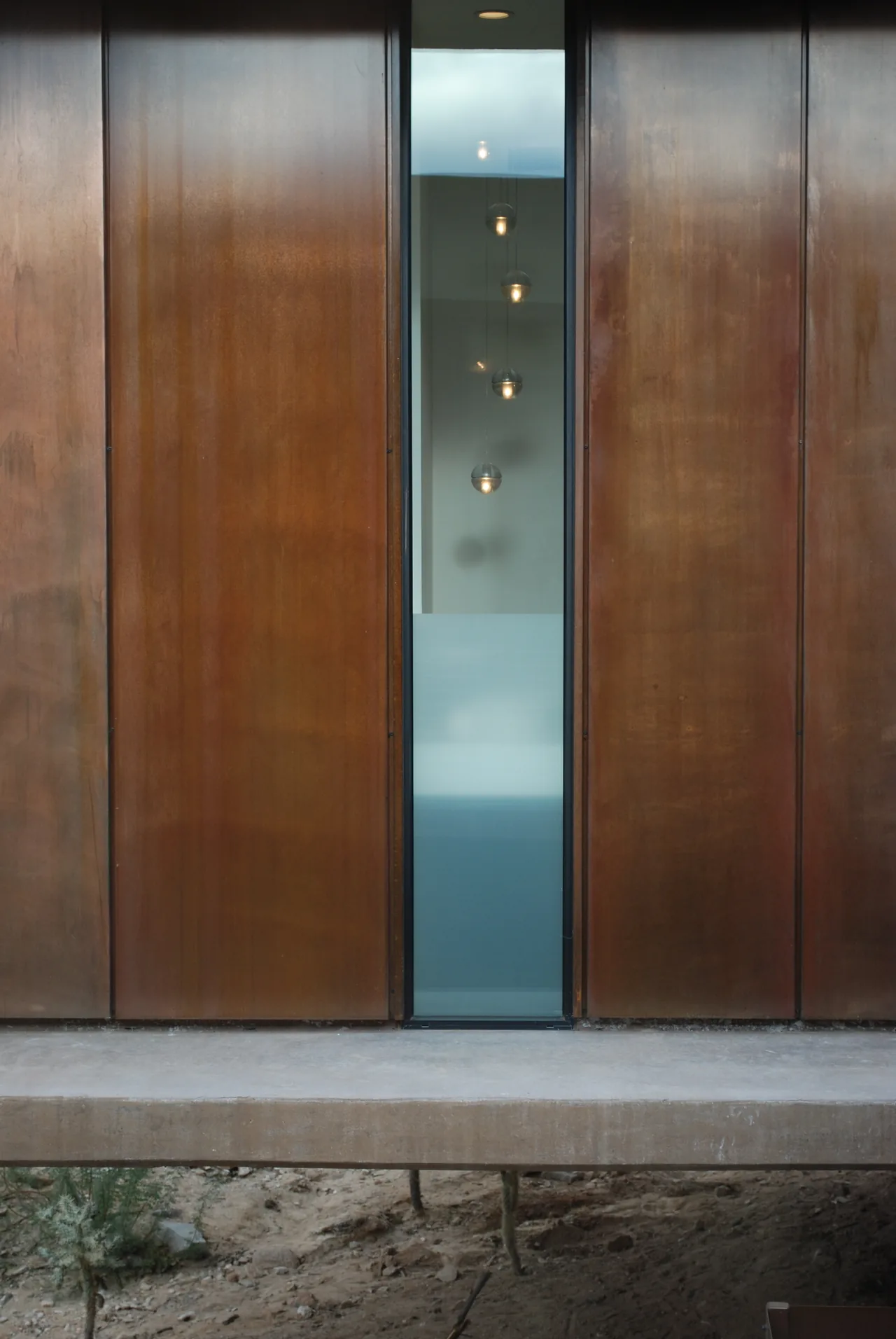 Window between weathered steel panels on an elevated structure over desert ground.