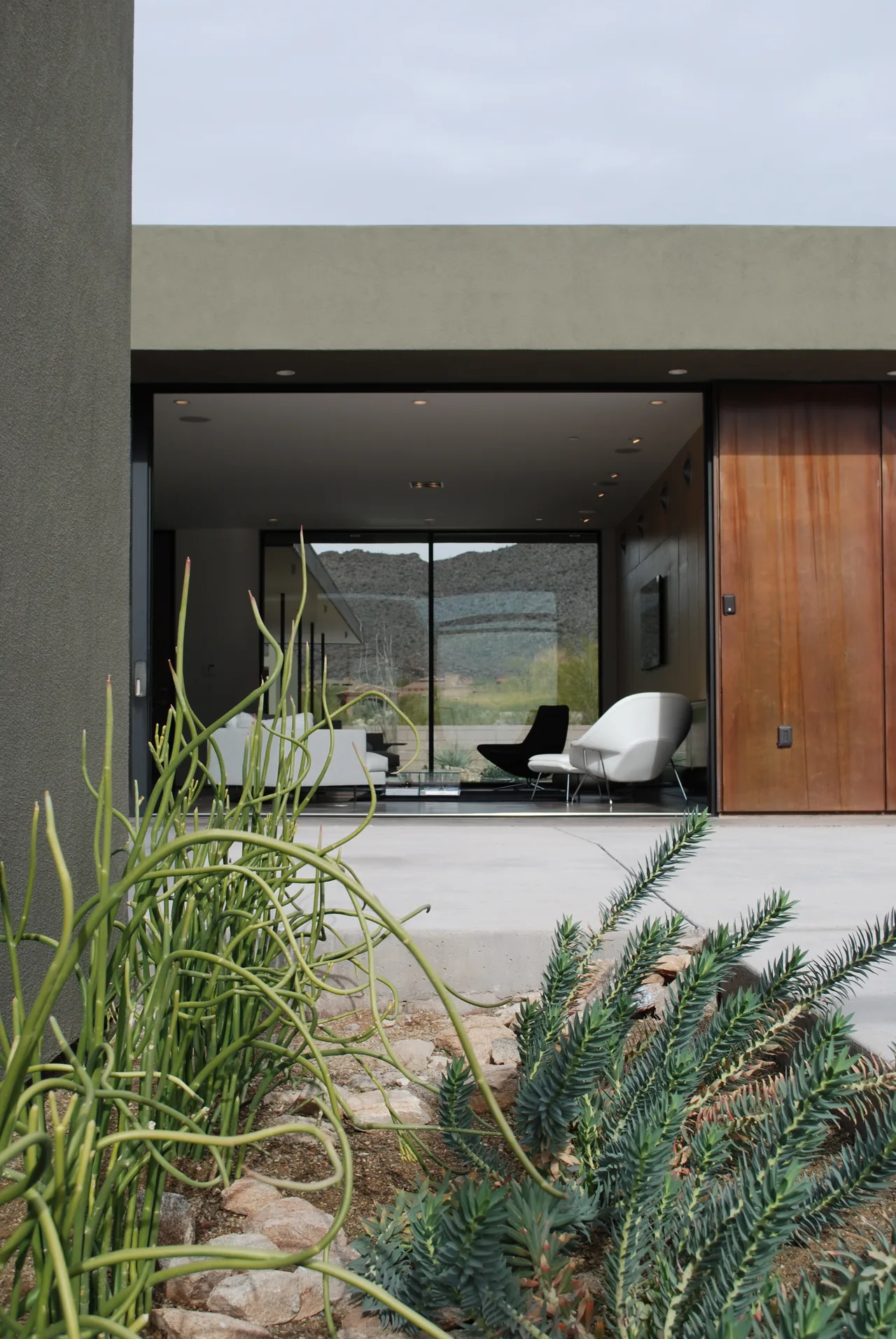 Modern home with an open wall revealing white and black chairs inside and desert landscaping outside.