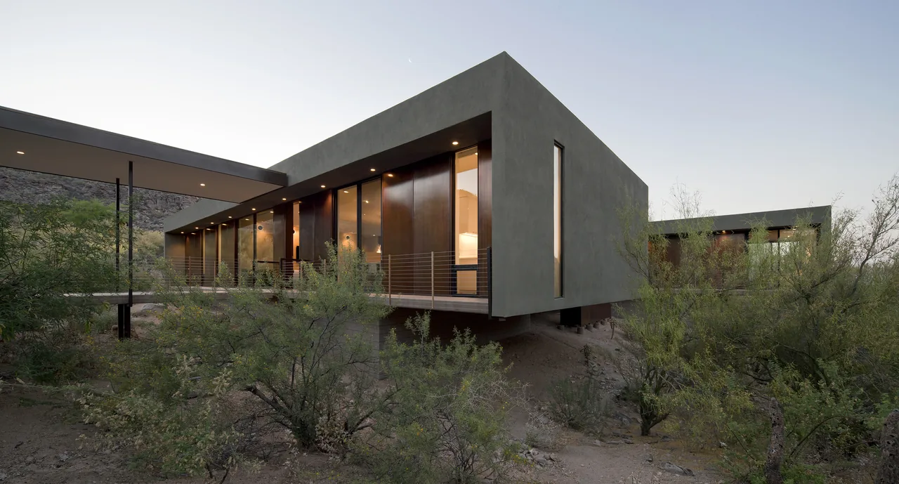 The IBARRA ROSANO project features a modern elevated home with large glass windows and a steel-post-supported outdoor walkway amid desert landscaping.