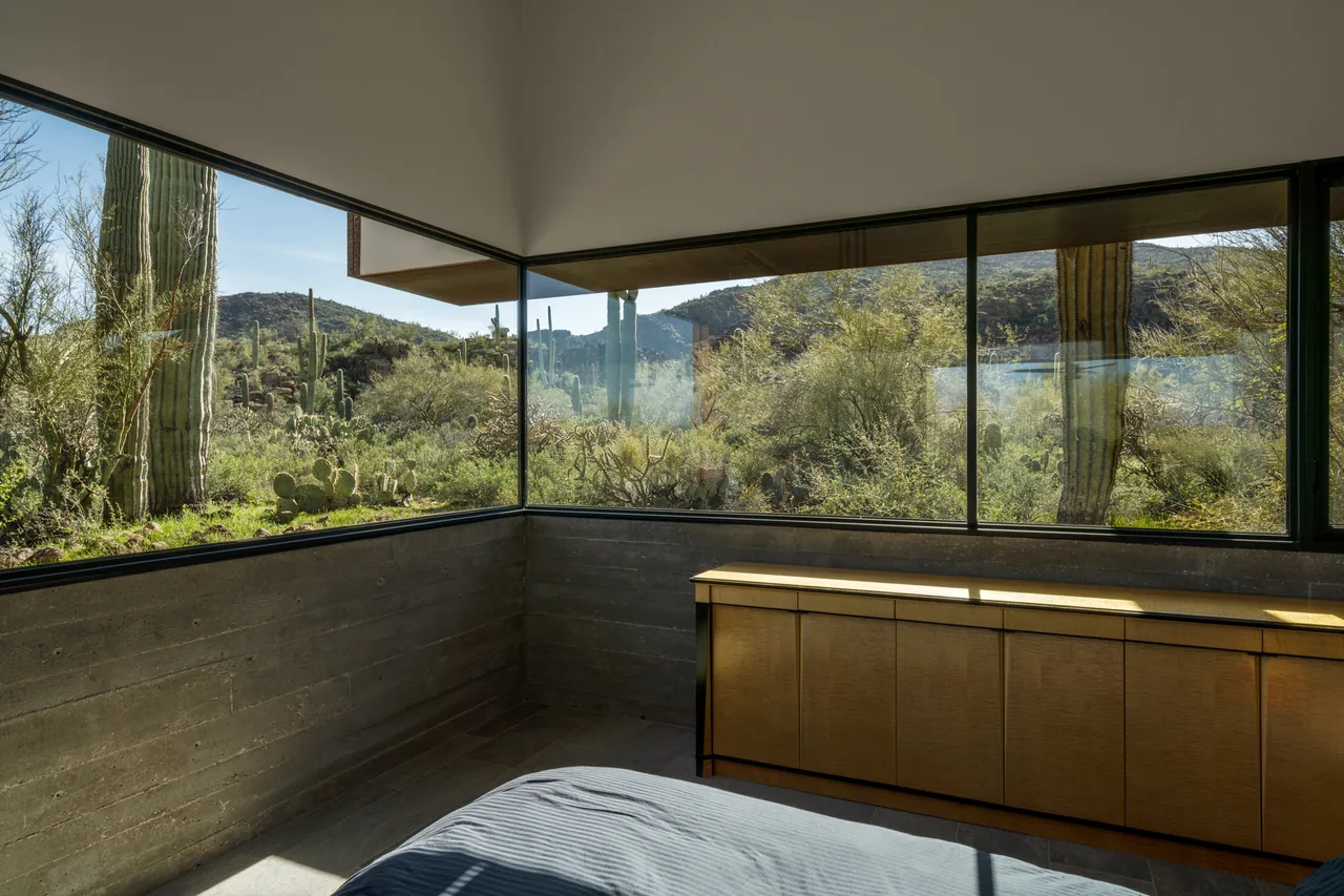 Bedroom corner in TS Falls West Residence features large windows with views of desert plants and mountains.