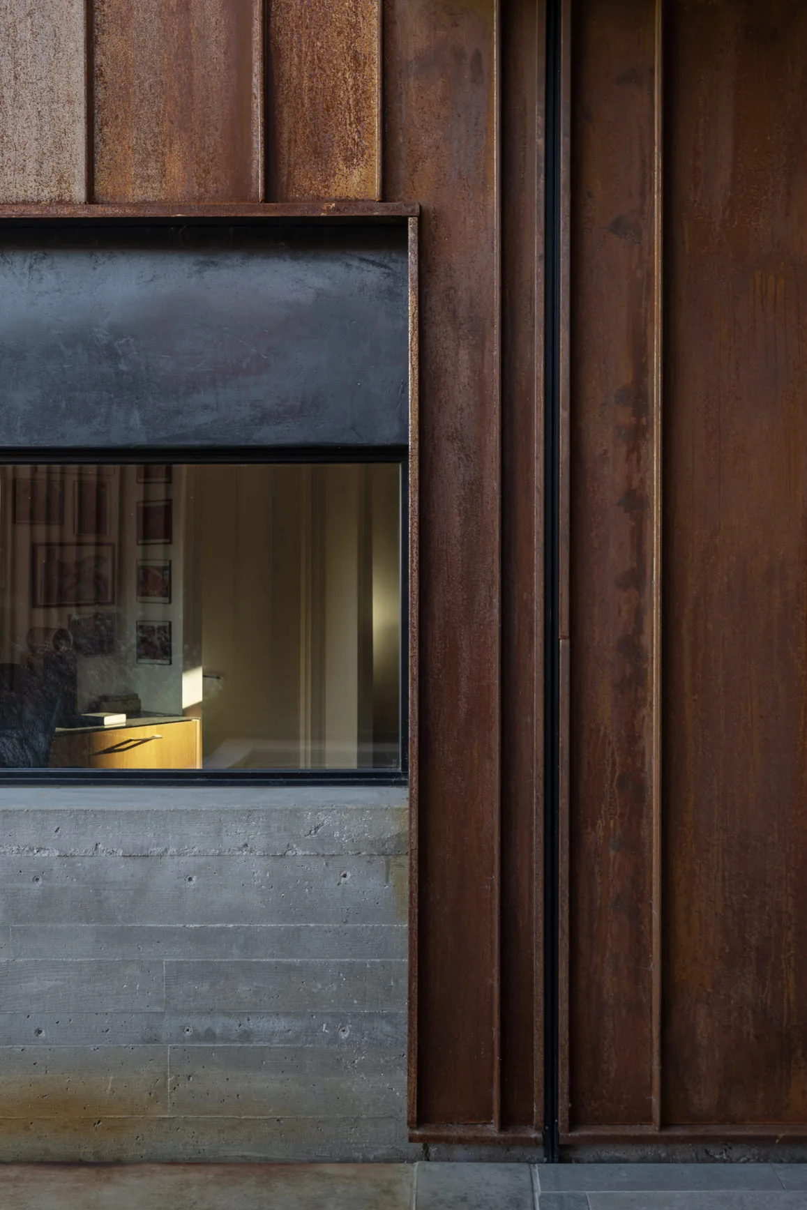 Exterior detail of rust-colored metal cladding with black window frame and concrete base at TS Falls West Residence.
