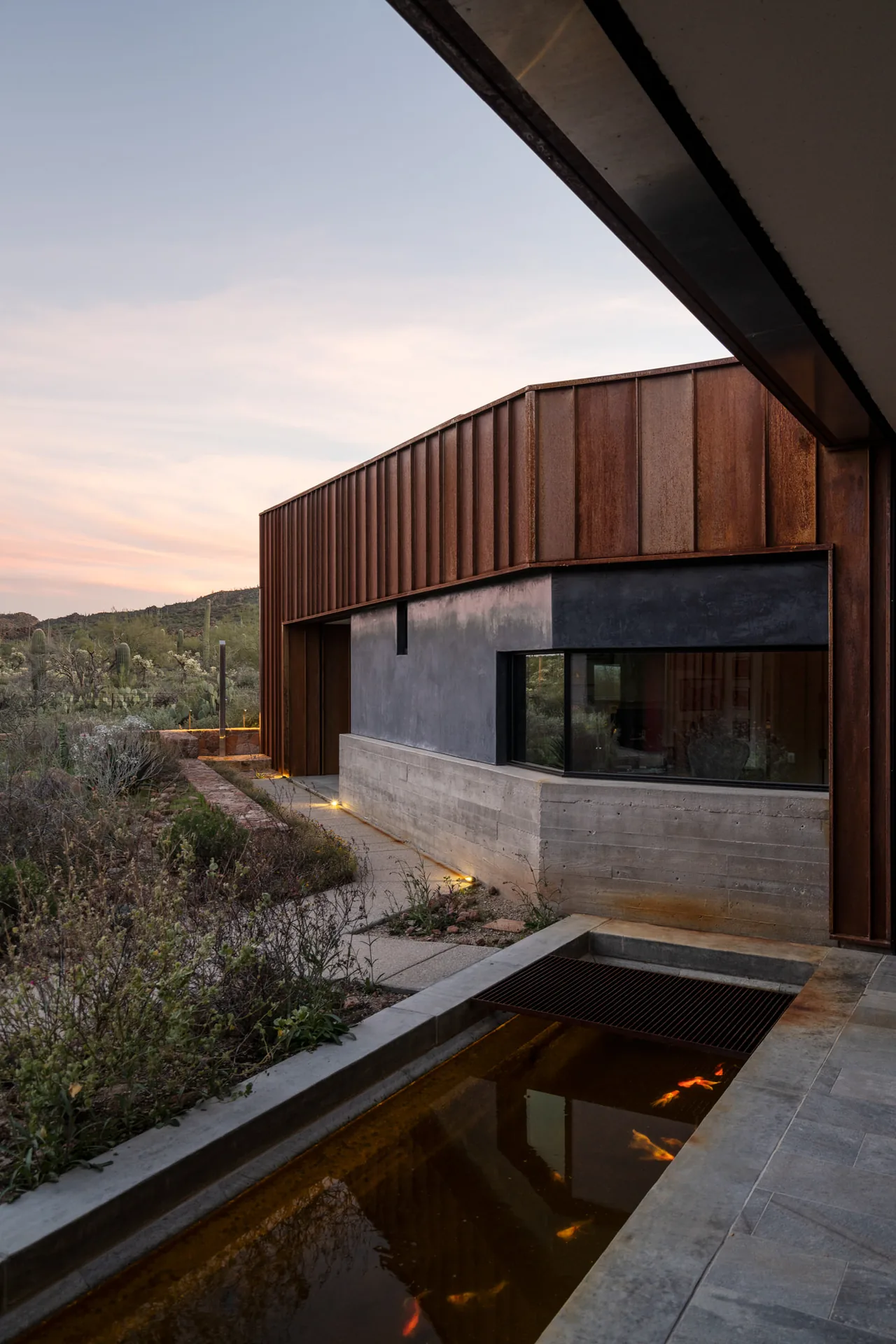 TS Falls West Residence showcases rust-colored metal cladding, concrete base, desert landscaping, and a reflective pool with fish.