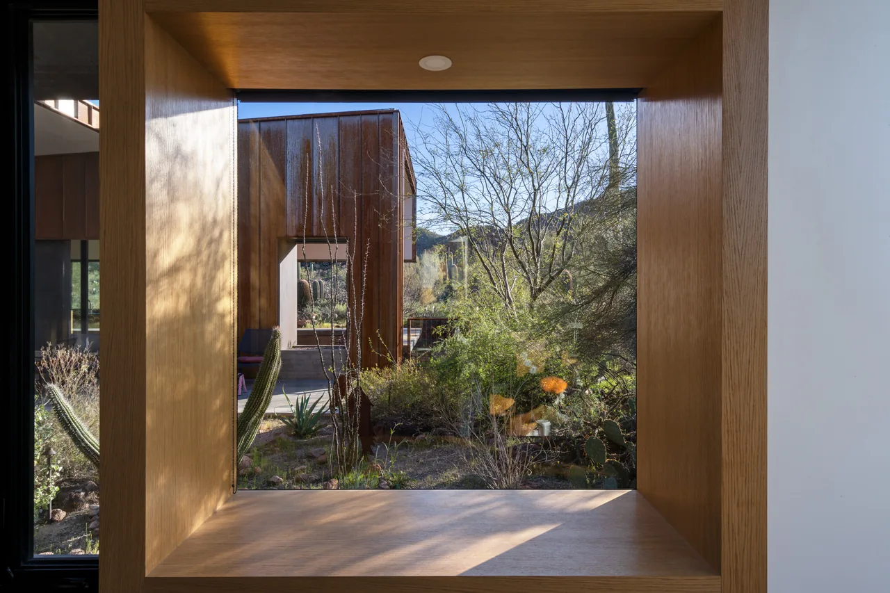 Square wood-framed window frames a view of desert plants and a rust-colored facade at TS Falls West Residence.