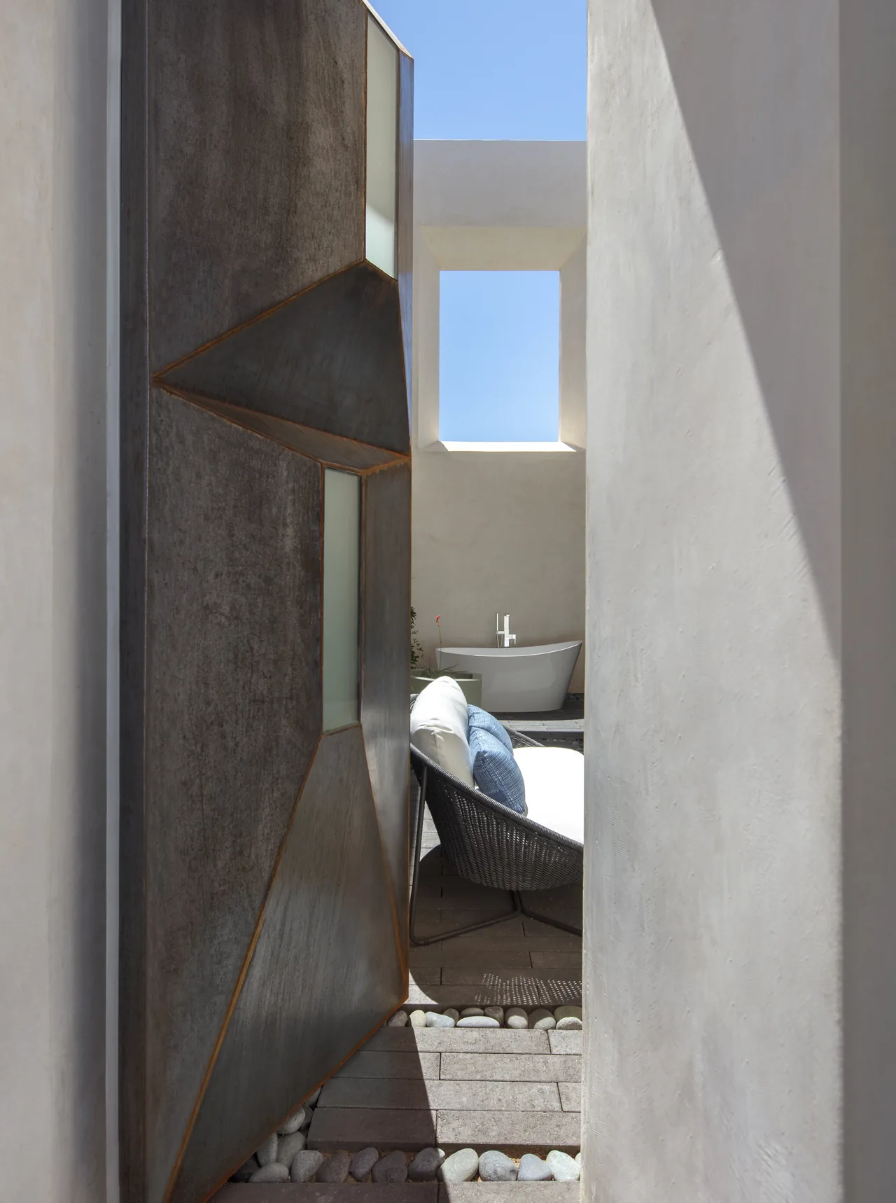 Casa Schneider features a courtyard with a geometric metal door, white stucco walls, and an outdoor lounge near a bathtub under a large square opening.