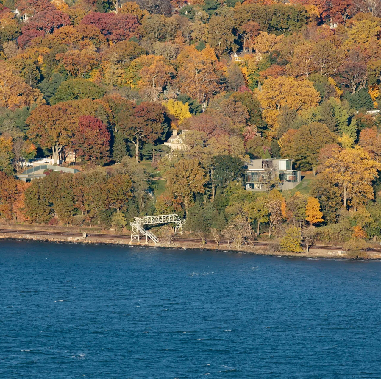 Horizon House sits on a sloped wooded site by the water, showing its modern form and cantilevered elements amid autumn foliage.