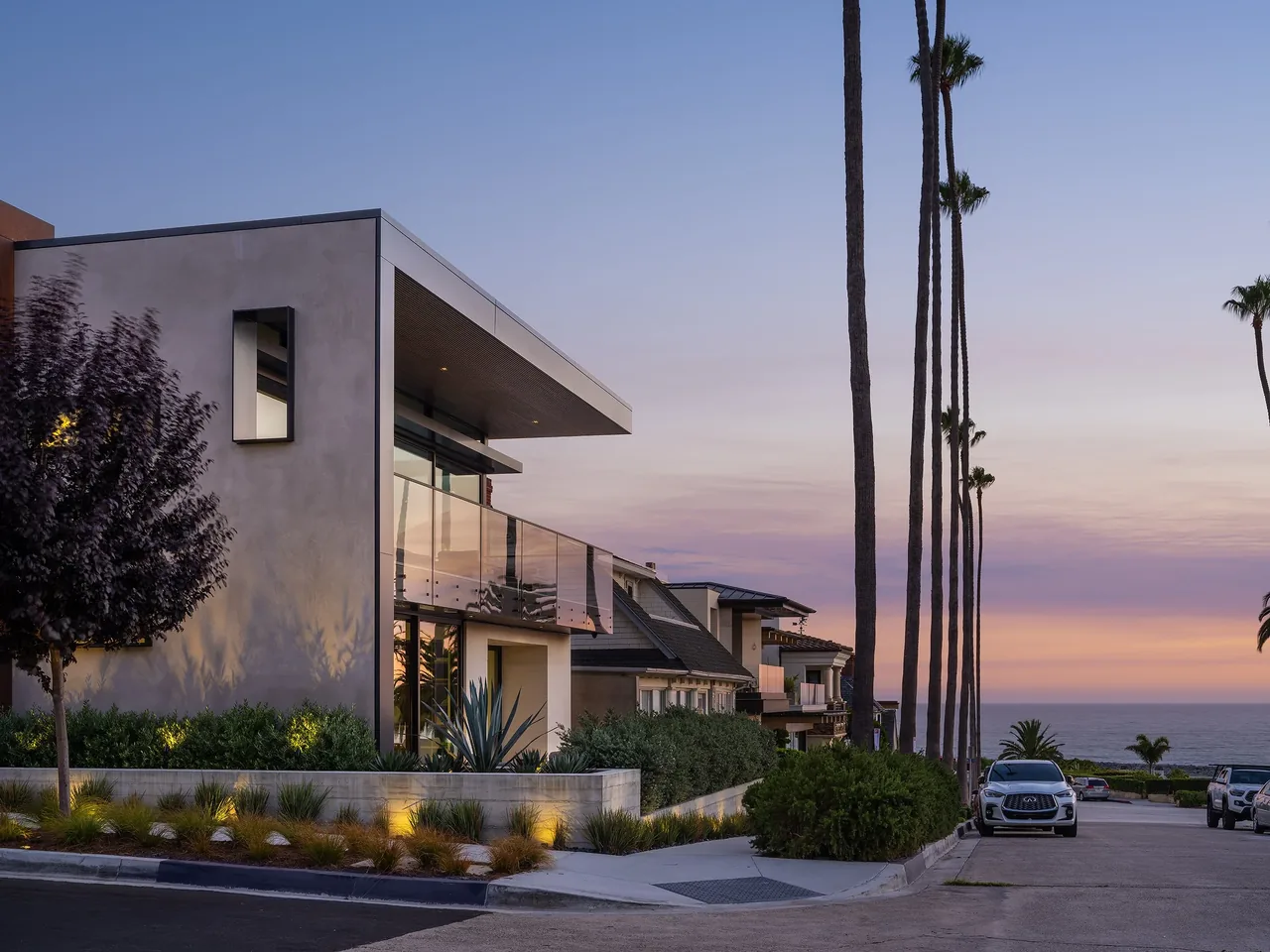 Goldenrod House features modern rectangular volumes with a glass balcony and landscaped sidewalk, set against a coastal sunset.