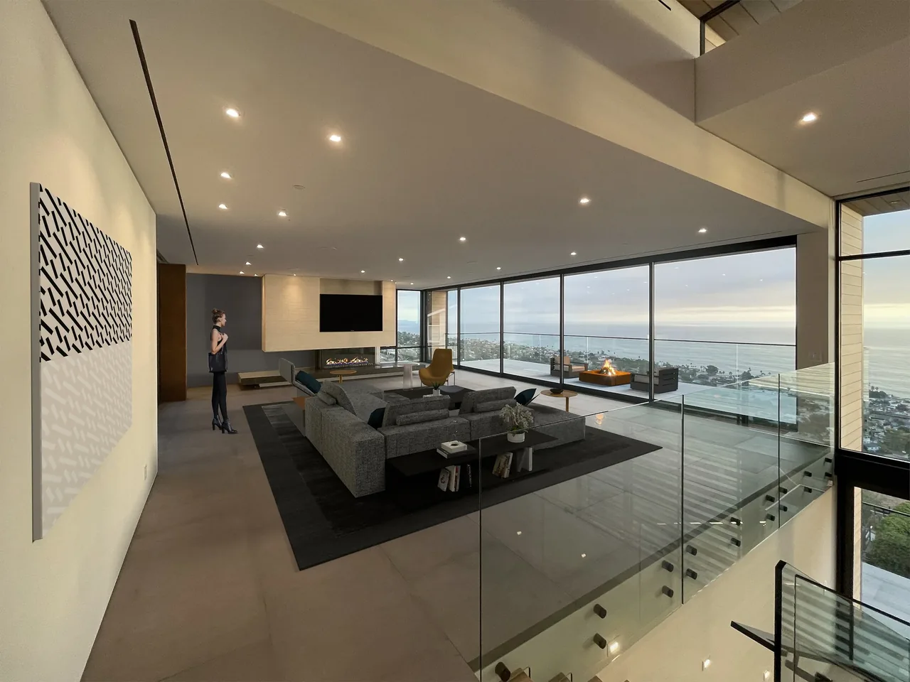 Wide living area with floor-to-ceiling glazing opens to a view of the ocean and hillside, featuring a modern fireplace and glass railing by the stairs.