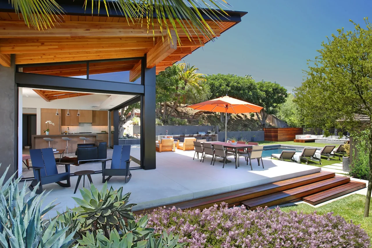 Modern pool house with an open kitchen, outdoor dining, and lounge seating under a wood-beamed roof.