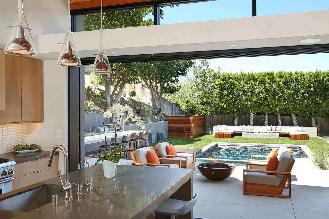 Indoor kitchen opens to a backyard with a pool, fire pit, and seating, showcasing sliding glass wall integration.