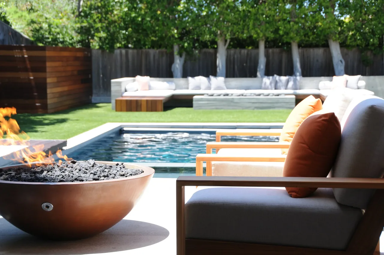 Poolside lounge chairs with orange pillows near a fire pit and long outdoor seating in the background.