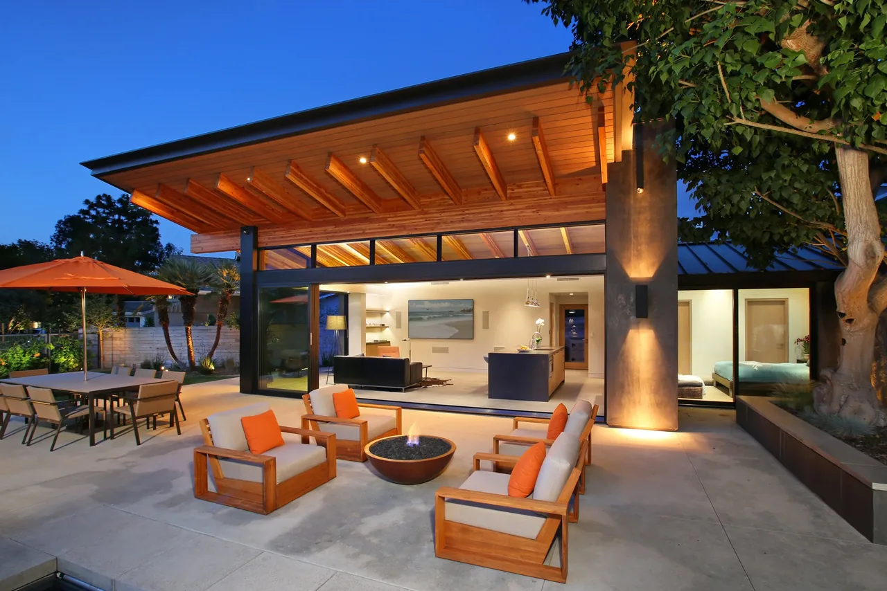 The VOLKOV POOL HOUSE features an outdoor lounge area with wooden chairs and a fire pit beside an open sliding wall to the interior living space.
