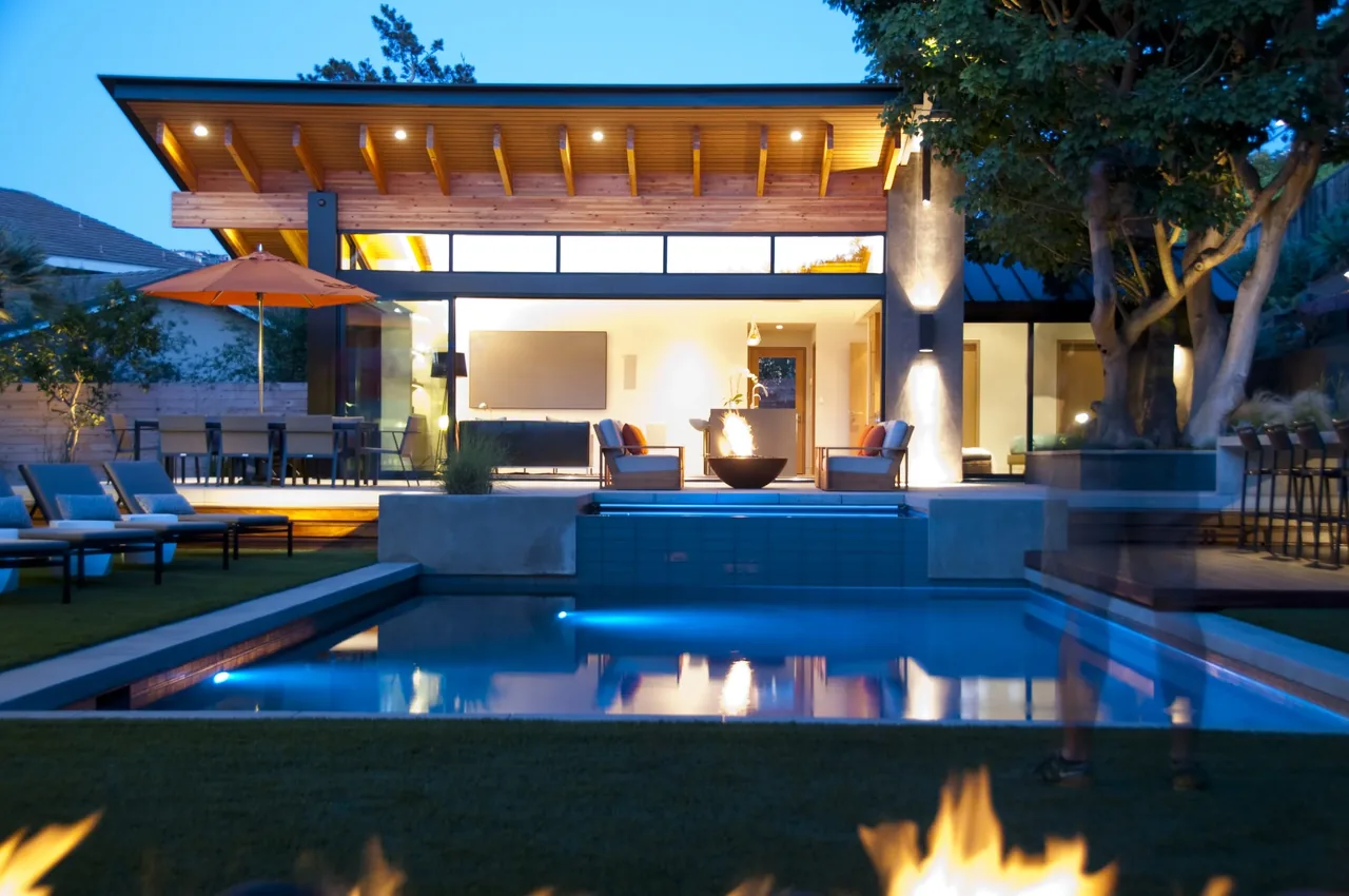 Modern pool house with open sliding wall, fire pit, and poolside seating under a flat roof with exposed beams.