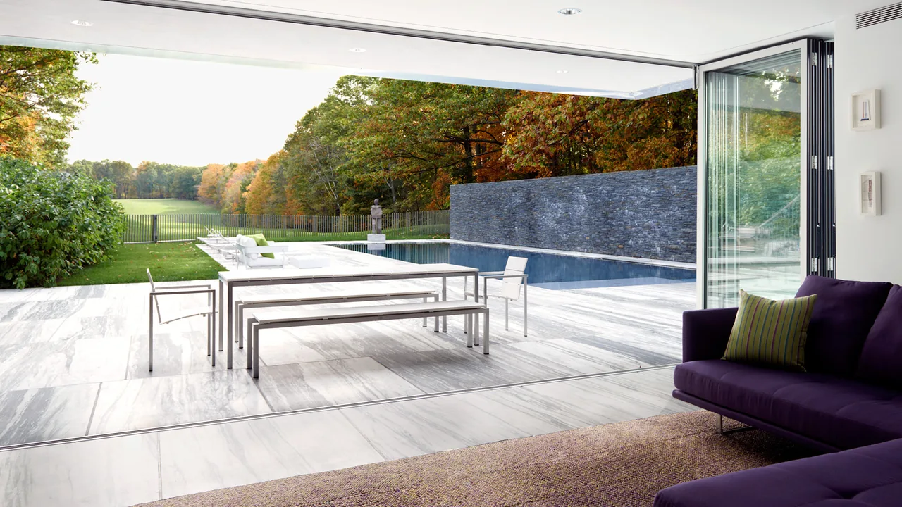 Living room opens seamlessly to an outdoor patio and pool area with a view of trees and lawn beyond.