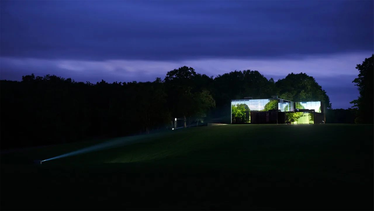 Dutchess County Residence by Allied Works Architecture features cubic form with illuminated glass panels reflecting surrounding nature at night.