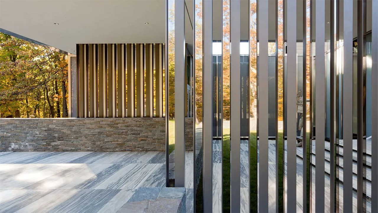 Exterior detail of paired vertical steel slats forming a privacy screen with stone wall and paved floor under an overhang.