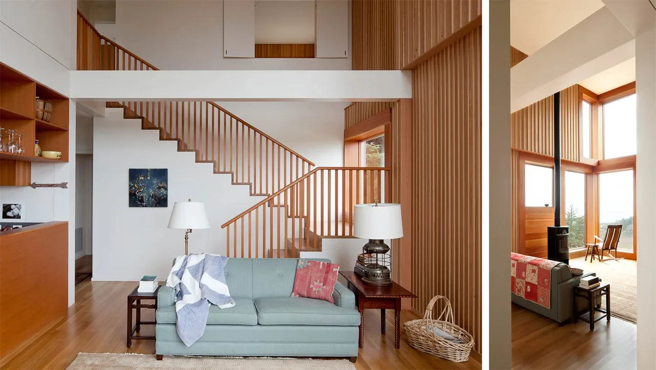 Living room with double-height ceiling, wood staircase, and large corner windows overlooking the outdoors.