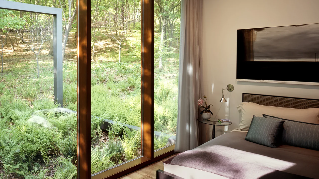Guest bedroom with floor-to-ceiling wood-framed windows facing a lush green forest setting.