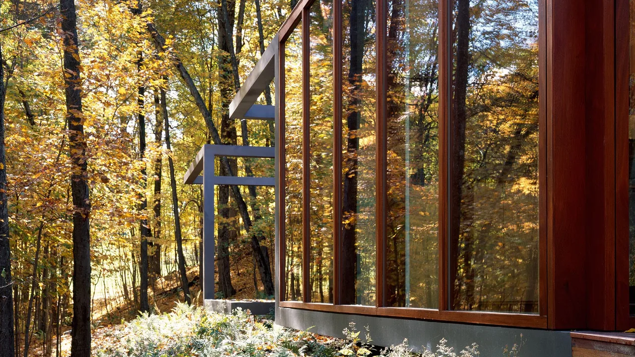 Guest house exterior with large glass panels framed in wood, reflecting fall foliage in a forested site.