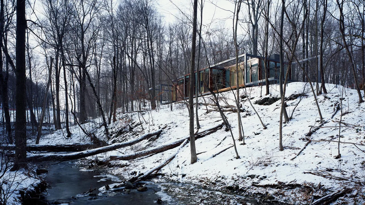 The Dutchess County Residence Guest House is situated on a snowy slope near a small creek, surrounded by bare winter trees.