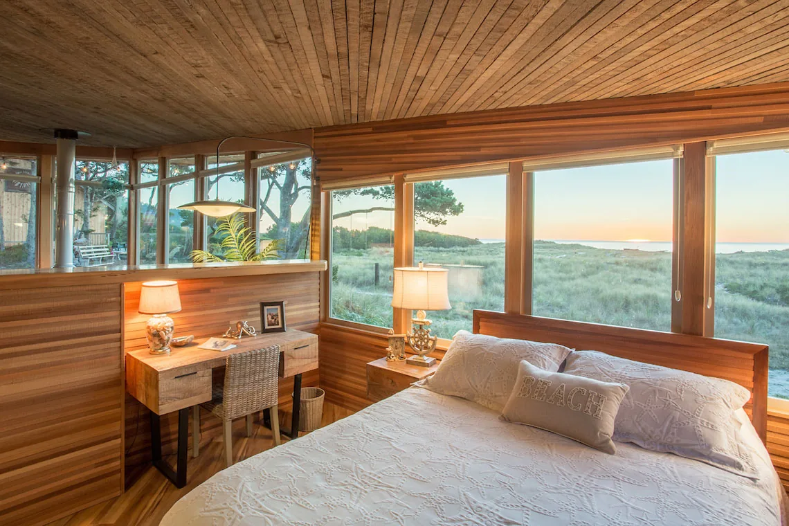 Bedroom interior with wood paneling and expansive coastal views through large windows at sunset.