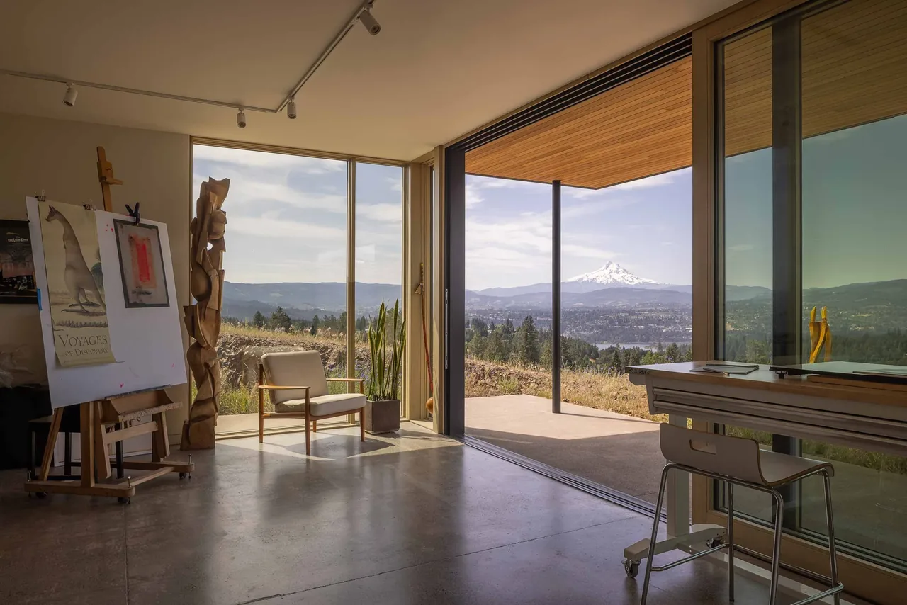 Interior view shows a light-filled room with glass walls framing a mountain landscape and an adjoining covered patio.