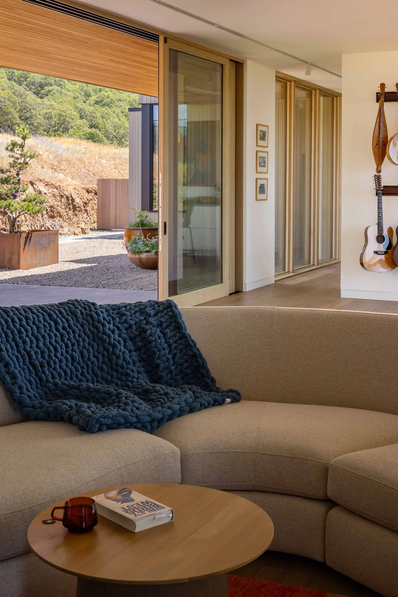 Cozy indoor living corner with curved sofa and large sliding glass doors opening to a natural gravel yard and hillside.