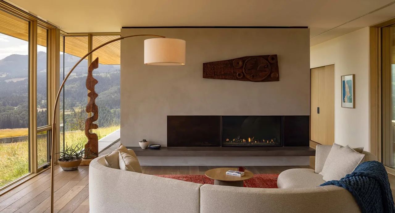 Living room of Bald Mountain Residence with a curved sofa, modern fireplace, and mountain backdrop visible through large windows.