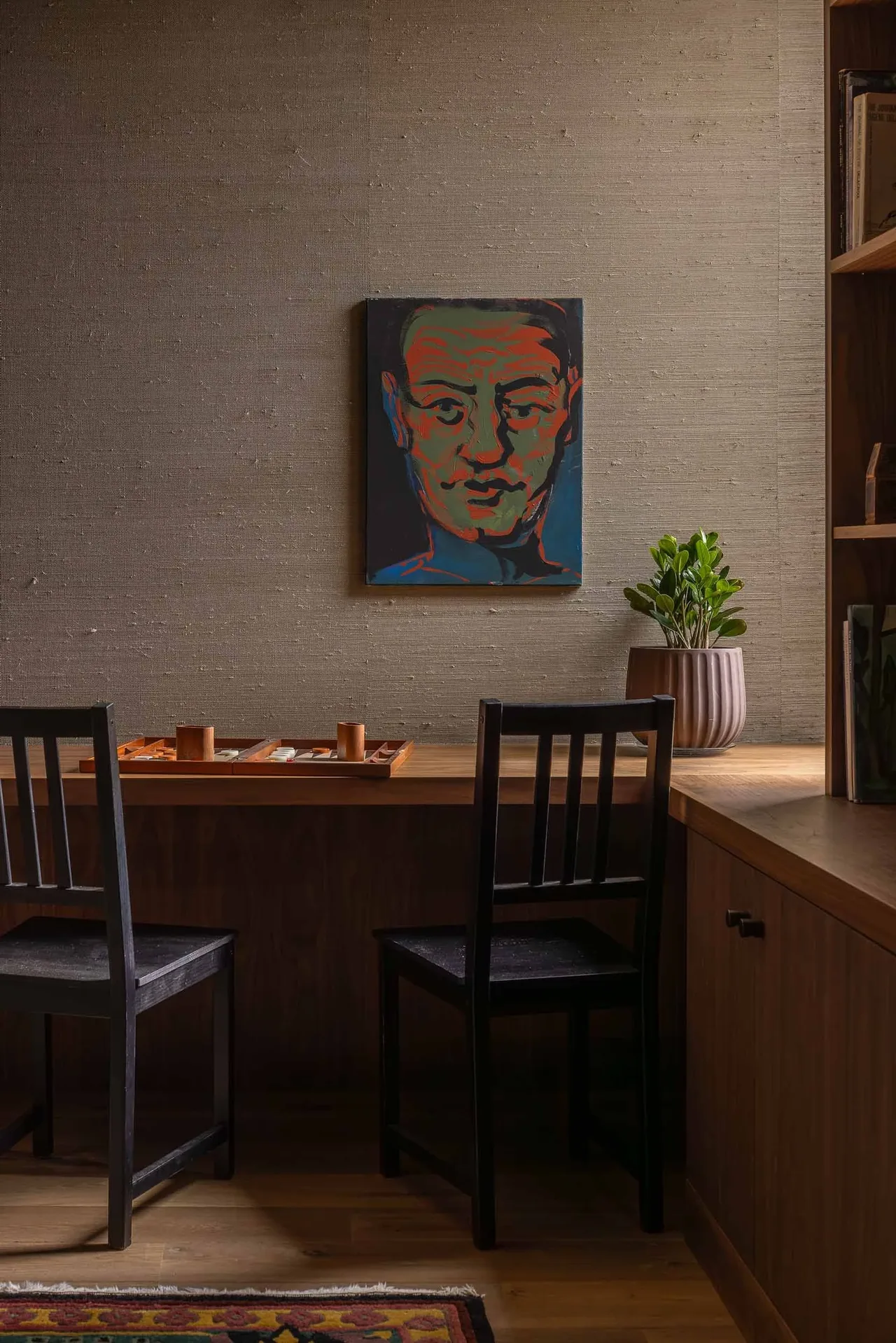 A study area features a wood desk with black chairs, a board game, and a potted plant under a portrait painting on a textured wall.